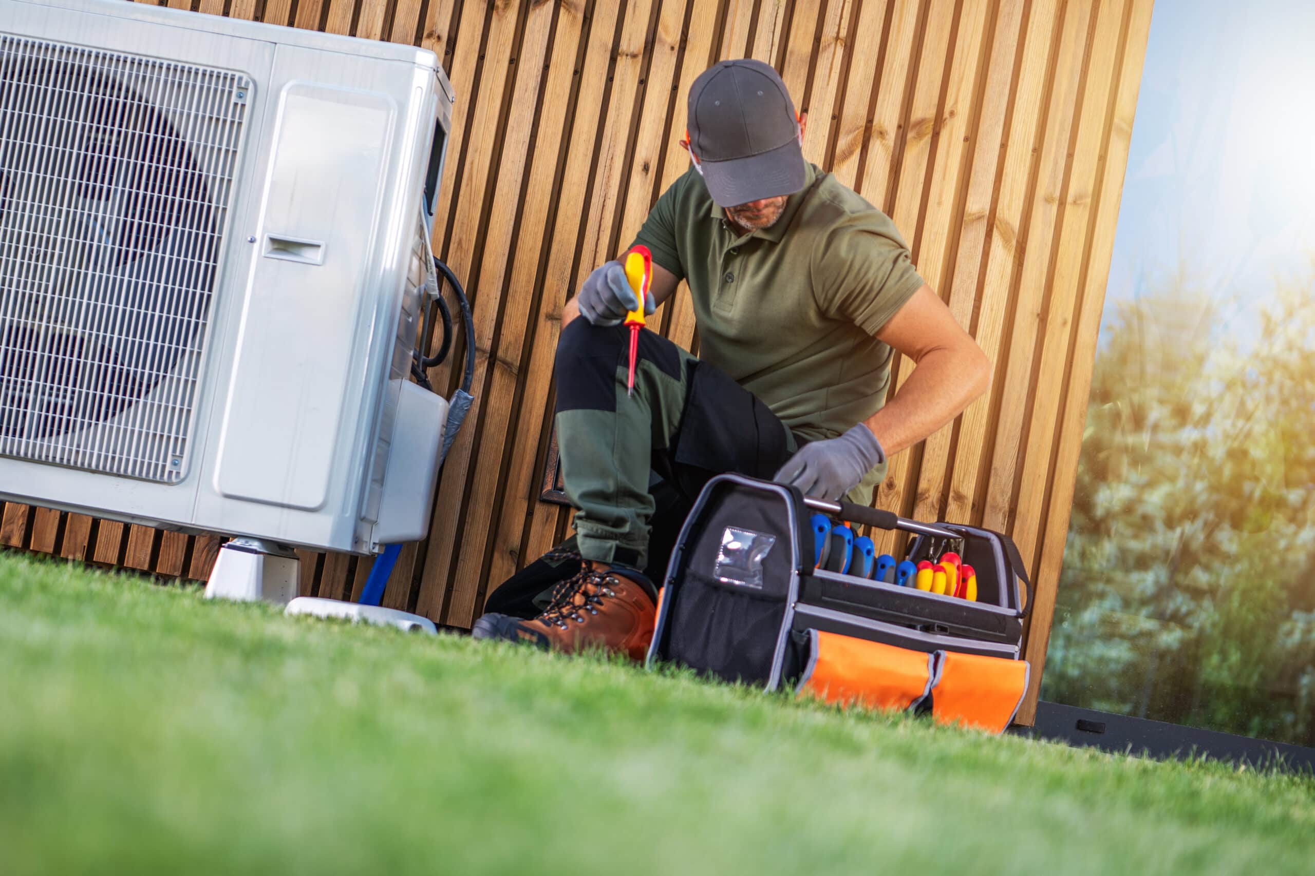A technician in a green shirt and gray cap sits on the grass, using tools from an open tool bag. An air conditioning unit is visible beside him, with a wooden wall in the background and natural light enhancing the scene.
