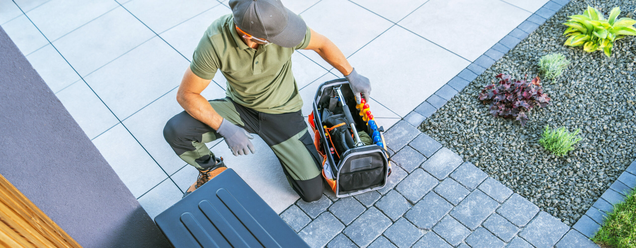 A person wearing a green shirt and gloves kneeling on a tiled outdoor surface while opening a tool bag. The surroundings include a landscaped area with stone borders and plants.