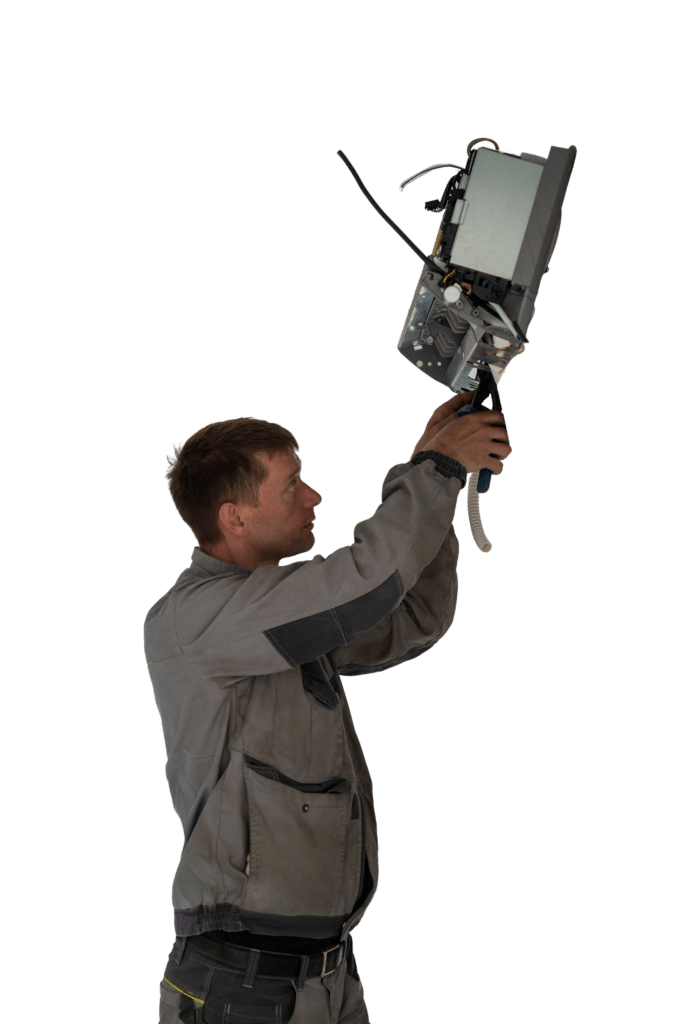 A technician wearing a gray jumpsuit is holding a telecommunications device above his head. He is focused on the equipment, which has several wires attached, set against a plain background.