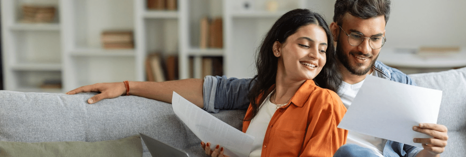 A man and a woman sitting on a gray couch, looking at documents in their hands. The woman is wearing an orange shirt and smiling, while the man, in glasses, is smiling and focusing on a sheet of paper. A bookshelf is visible in the background.