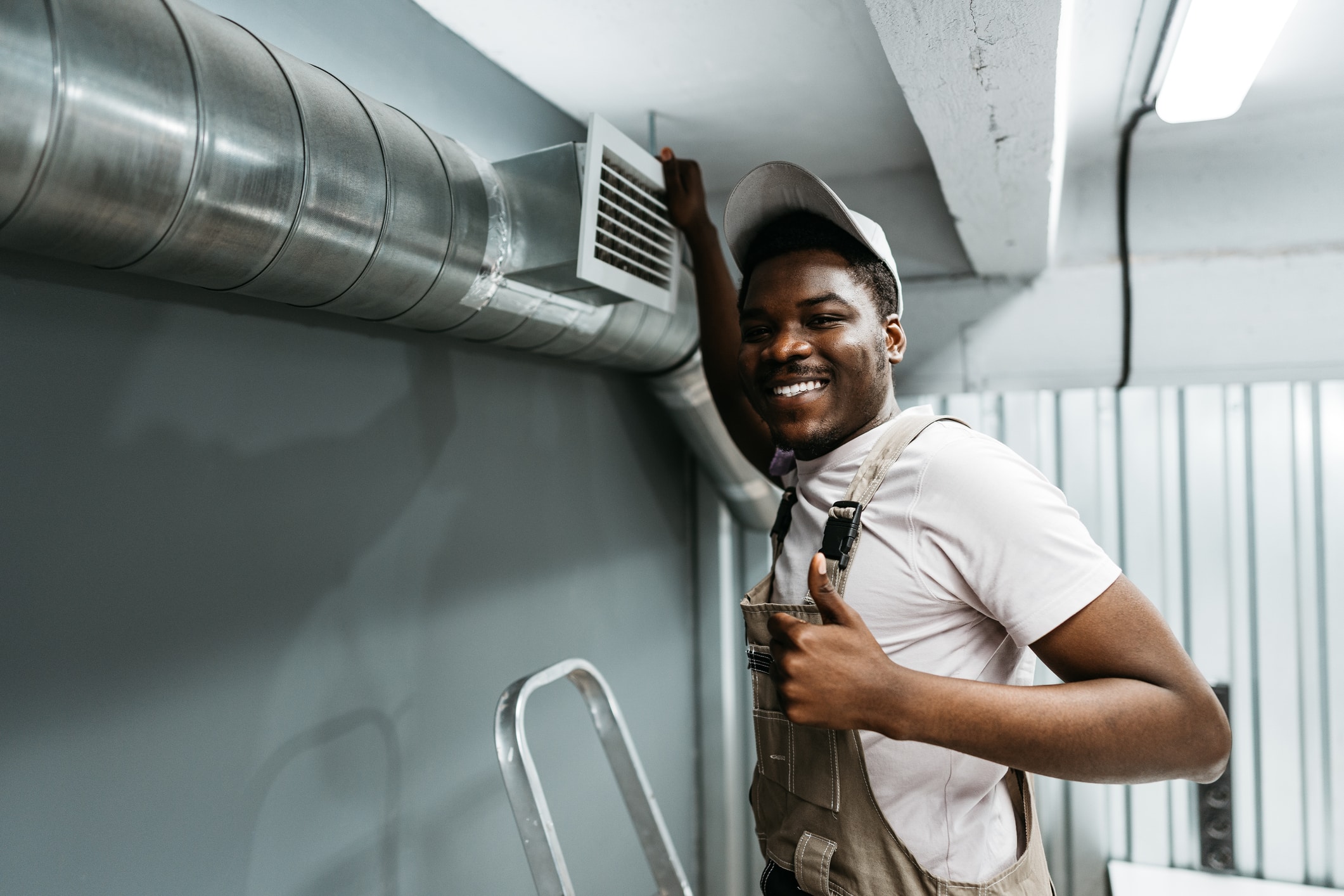 A smiling young man wearing a white shirt and brown overalls stands next to a metal air vent on a wall. He gives a thumbs-up gesture with his right hand, while leaning against a ladder in a well-lit, industrial-style space.