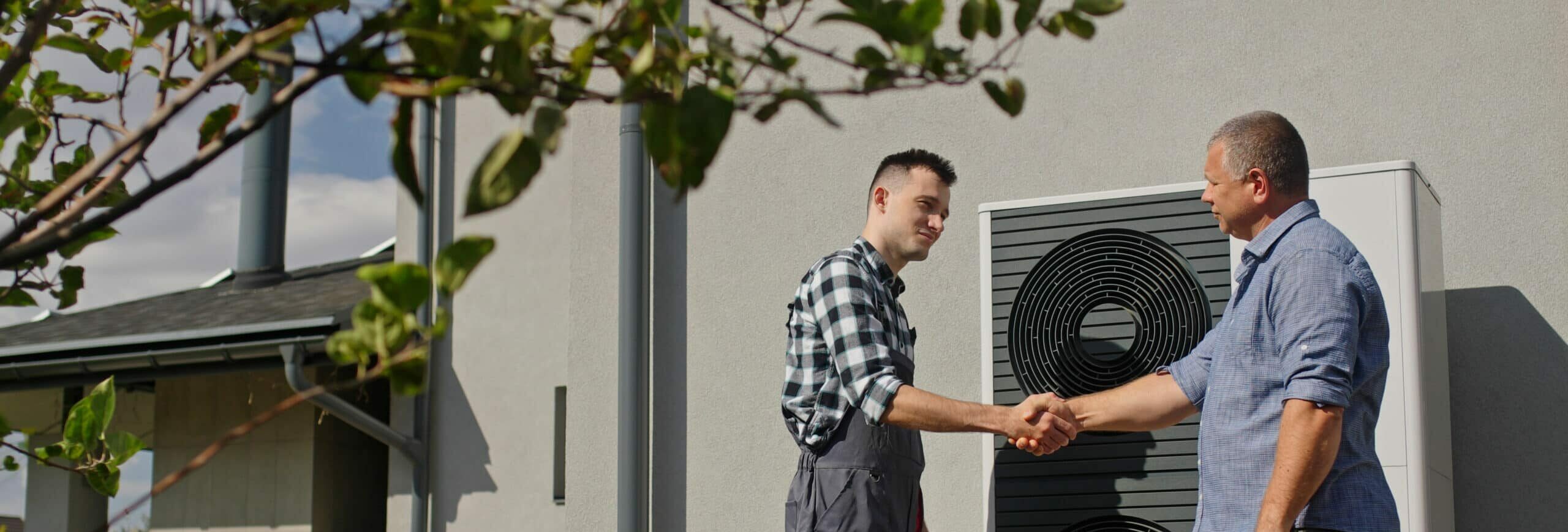 Two men shaking hands in front of a modern building exterior with an air conditioning unit on the wall. One man is wearing a black and white checked shirt and the other is in a blue shirt. Green leaves are visible in the foreground.