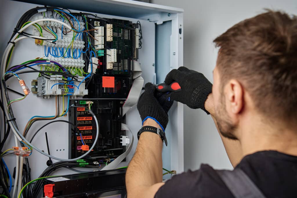 A technician wearing black gloves works on an electrical control panel. The panel is filled with various wires, circuit boards, and components. The technician adjusts a connection using a screwdriver, focused on the task.