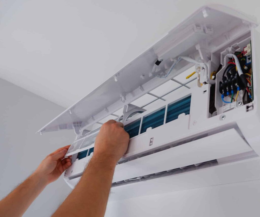 A person using both hands to open a mounted air conditioning unit on a wall. The air conditioner is partially disassembled, revealing internal components such as wires and a filter. The background is a plain wall in a well-lit room.