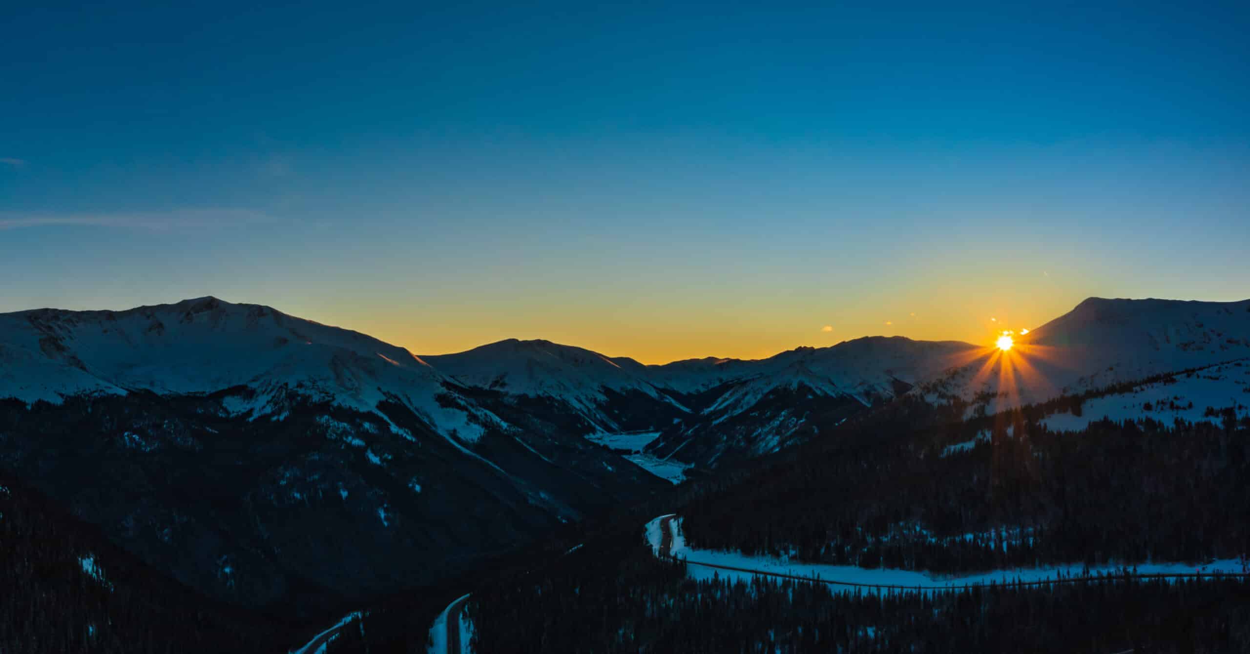 A panoramic view of snow-covered mountains at sunrise. The sky transitions from deep blue to warm orange near the horizon, with the sun peeking out from behind the peaks. The landscape features valleys and a winding river in the foreground.