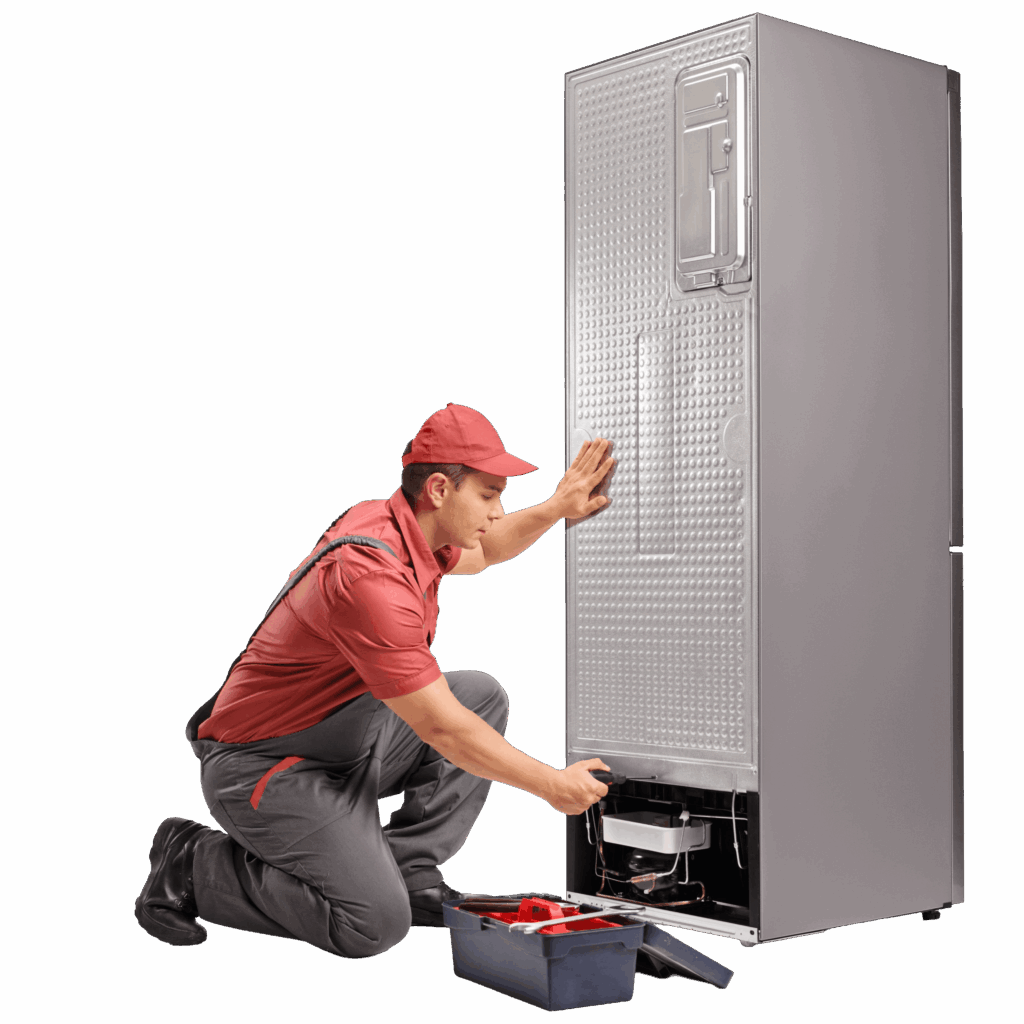 A technician in a red shirt and cap is kneeling beside a refrigerator. He is inspecting the bottom part of the unit and is holding a tool from an open toolbox placed near him. The refrigerator has a textured silver surface.