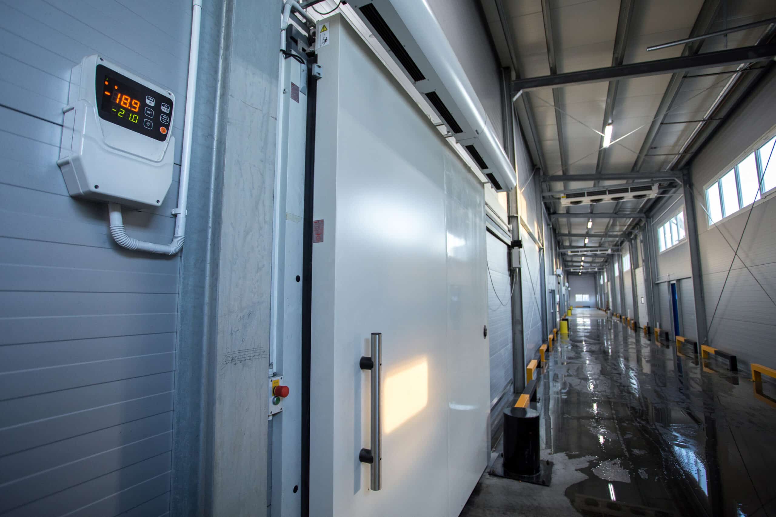 A large commercial freezer door in a warehouse, equipped with a digital temperature display showing -18.9°C. The corridor is well-lit with a polished floor and yellow loading docks along the wall.