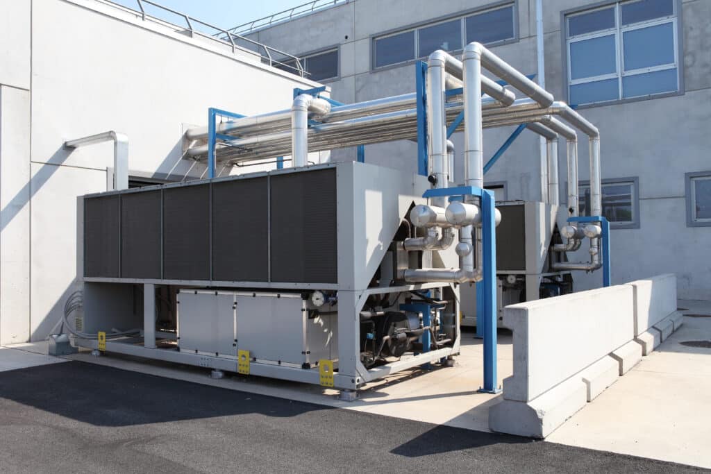 Two large industrial chillers mounted outside a modern building, featuring metallic frames and multiple connected pipes. The chillers have dark grids and are situated on a concrete surface with a dividing wall in front.