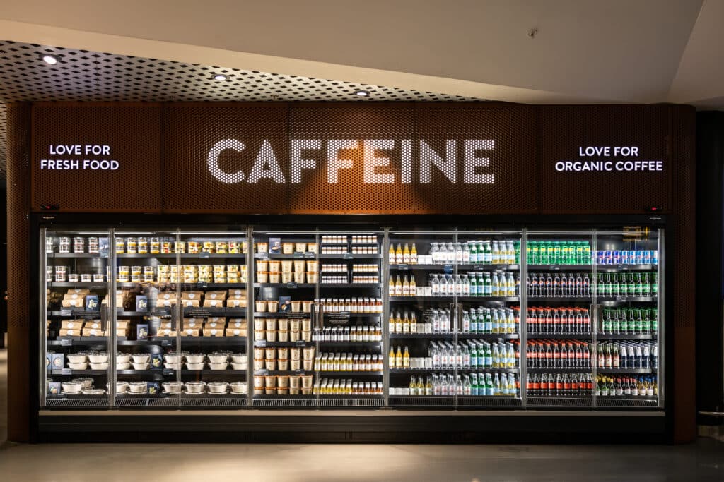 A display case labeled "CAFFEINE" showcasing a variety of bottled beverages, snacks, and packaged food items. Shelves are well-organized with products including organic coffee and soft drinks. Two signs state "LOVE FOR FRESH FOOD" and "LOVE FOR ORGANIC COFFEE."