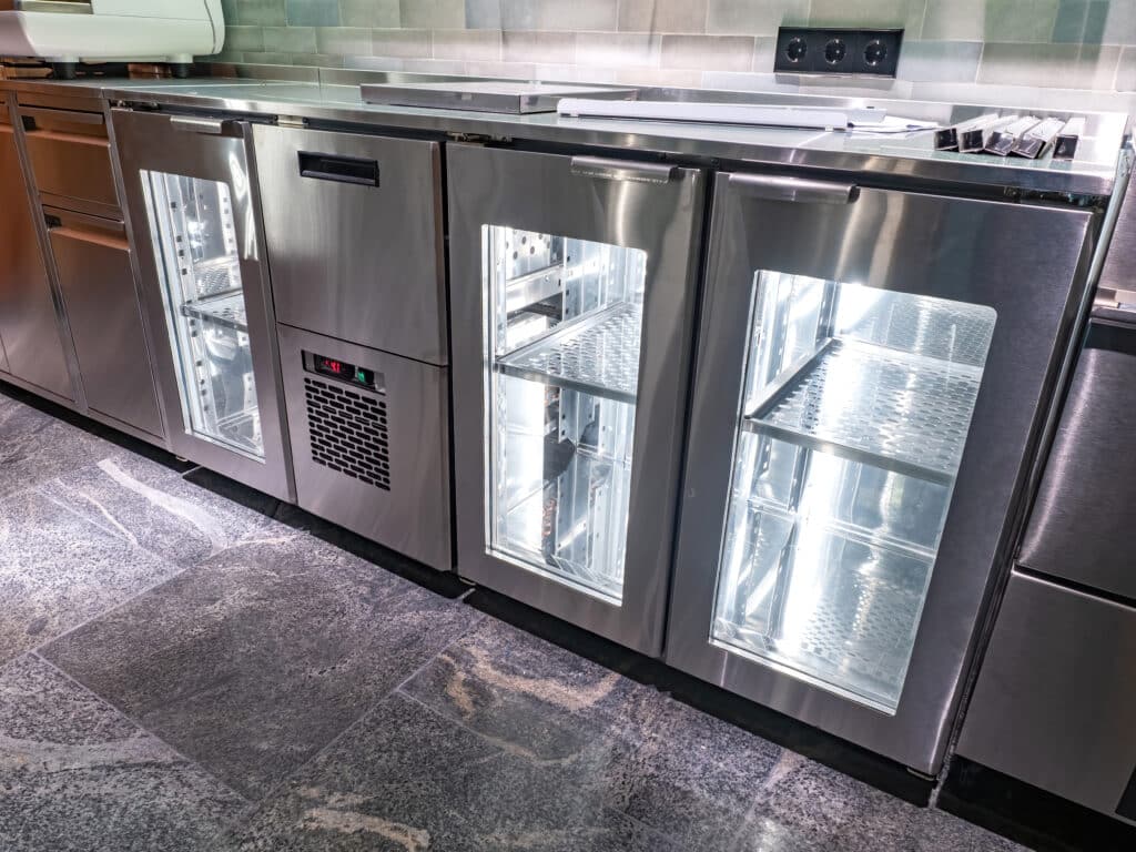 A row of three modern stainless steel refrigerators with glass doors in a kitchen. The interior of the refrigerators is illuminated, showcasing empty shelves. The floor is made of dark stone tiles, and the kitchen has a contemporary design.
