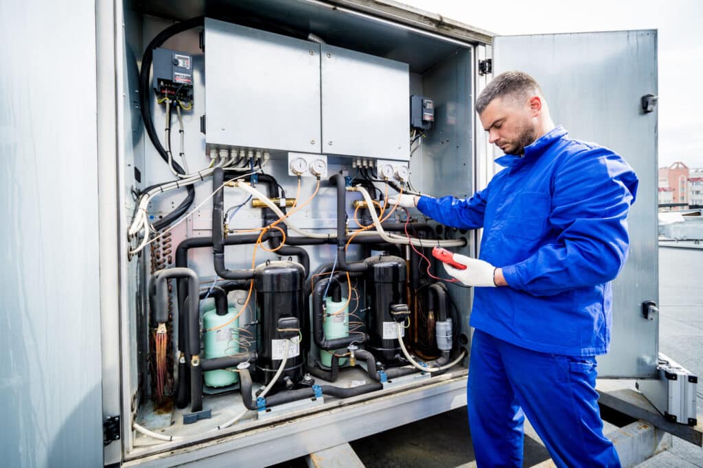 A technician in a blue uniform inspecting the interior of a refrigeration unit outdoors. He is holding a tool and standing next to various components, including compressors and pipes, inside a gray metal enclosure.