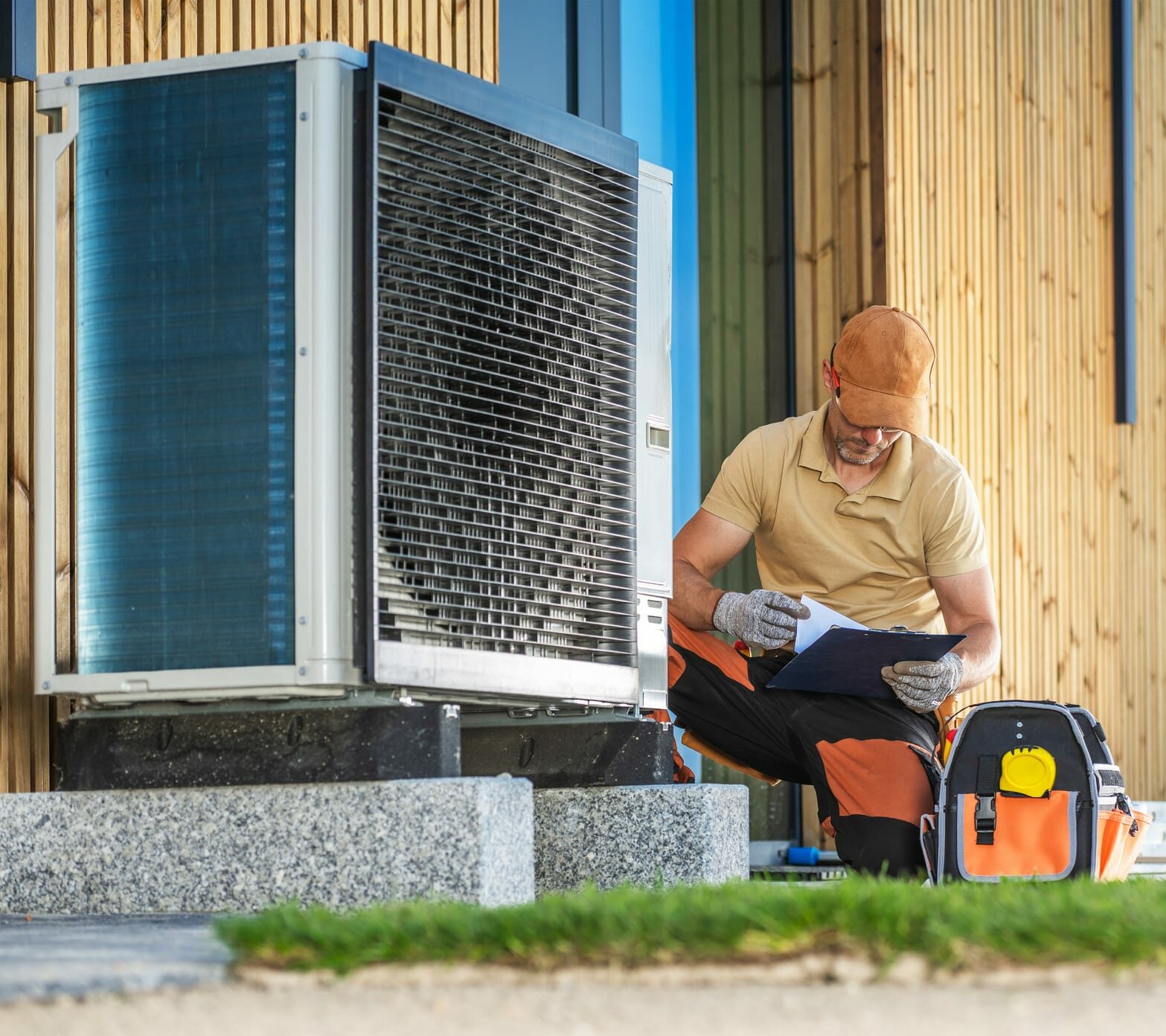 A technician in a cap is kneeling beside an air conditioning unit outside a building, taking notes on a clipboard. The technician is wearing gloves and has a tool bag nearby, with a backdrop of wooden paneling and blue walls.