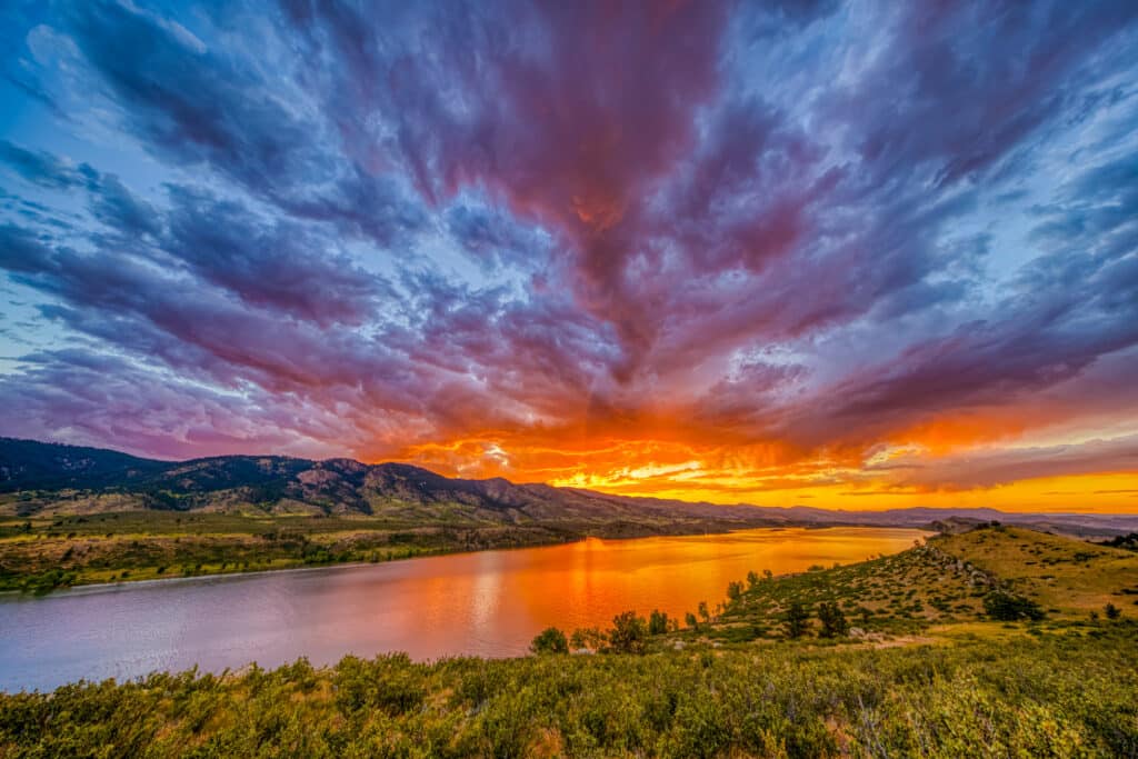 A panoramic view of a lake surrounded by rolling hills under a vibrant sunset sky. The clouds are illuminated in shades of orange, purple, and blue, reflecting on the calm water surface, highlighting the natural landscape's beauty.