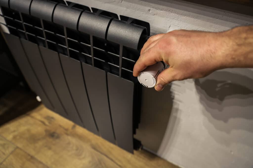 A hand adjusting the control knob of a modern radiator. The radiator has a sleek, black design with multiple vertical slats. The wall behind the radiator is light-colored, and the floor features wooden planks.