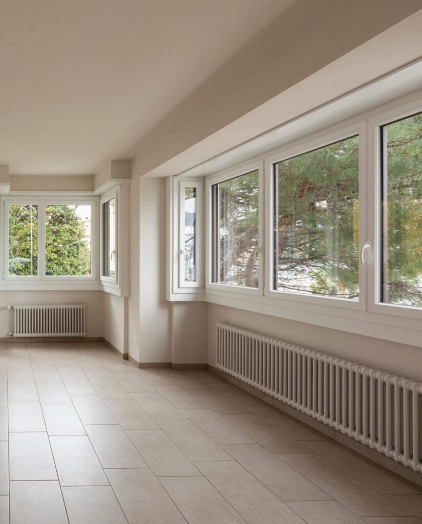 An empty room with light-colored walls and large windows allowing natural light. The floor is tiled, and there are radiators beneath the windows. Outside the windows, greenery is visible.