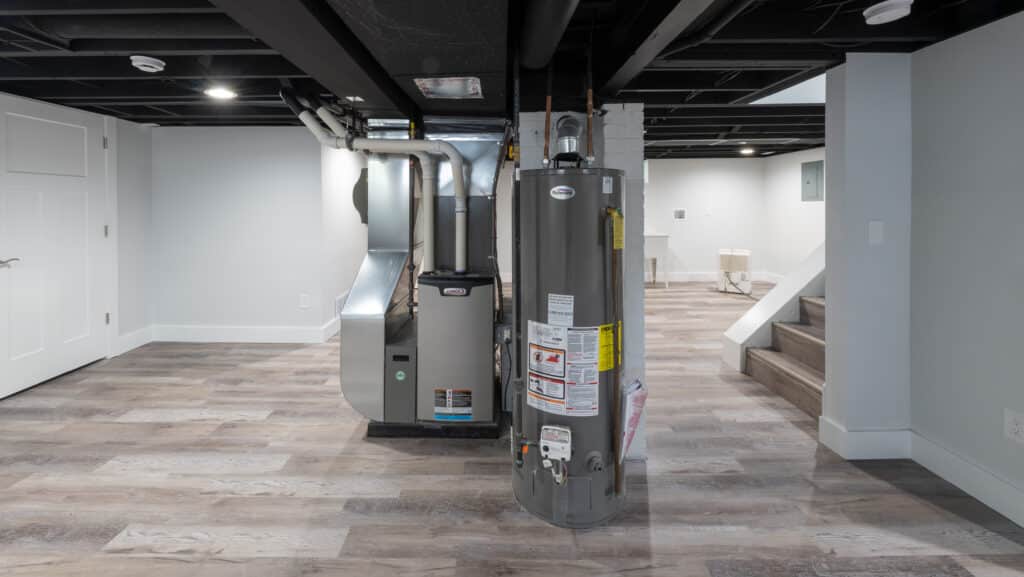 Interior view of a basement showing a heating system on the left and a water heater on the right. The space features light wooden flooring, white walls, and a staircase in the background leading to a different level. Recessed lighting is installed in the ceiling.