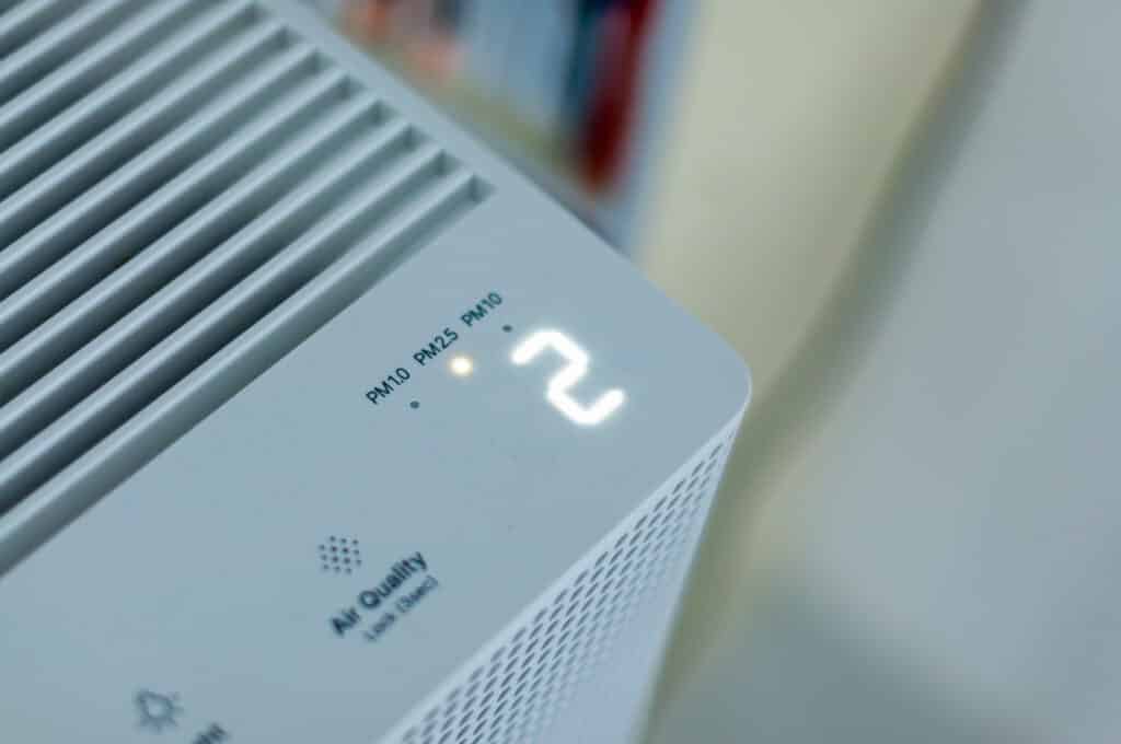 Close-up of an air quality monitor displaying numeric values. The screen shows indicators for PM10, PM2.5, and PM0, with a highlighted digit '2.' The device features a white design with ventilation slits and is positioned near blurred books in the background.