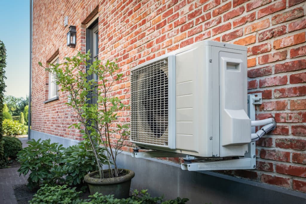 An outdoor air conditioning unit mounted on a brick wall next to a green shrub in a planter. The building is made of red brick with a wooden door and a light fixture visible. The scene is well-lit and shows a landscaped area.