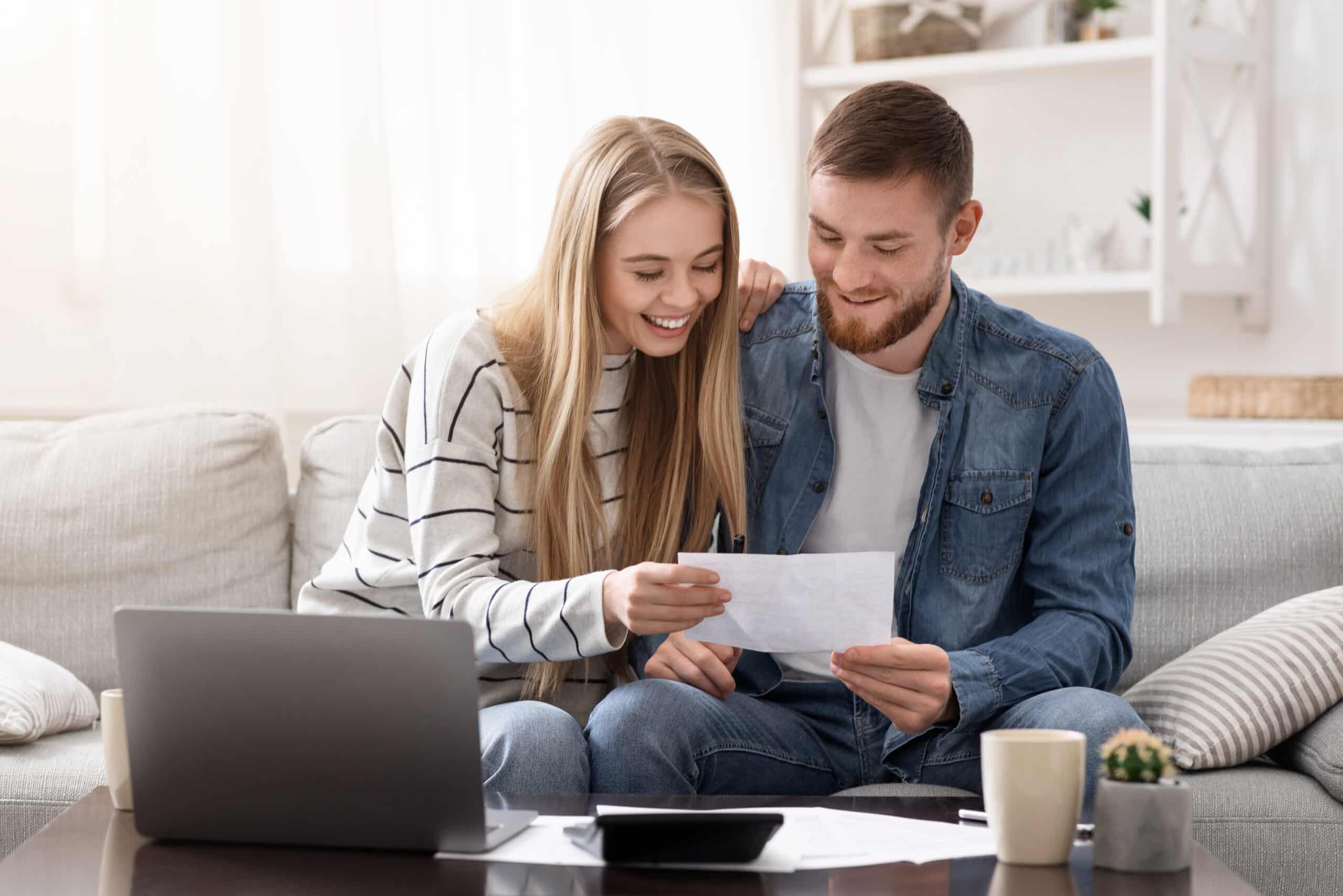 A young couple sitting on a couch, looking at a document together. The woman, with long blonde hair, smiles while holding the paper. The man, with short brown hair, leans in closely. A laptop and coffee cups are on the table in front of them.