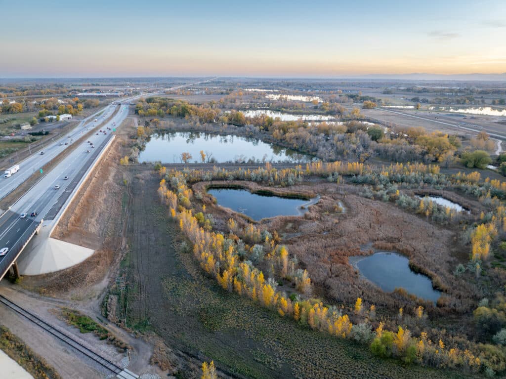An aerial view of a highway alongside a landscape featuring several ponds and patches of trees. The scenery includes a variety of greenery and autumn foliage, with a clear sky in the background, showing an expansive rural setting.