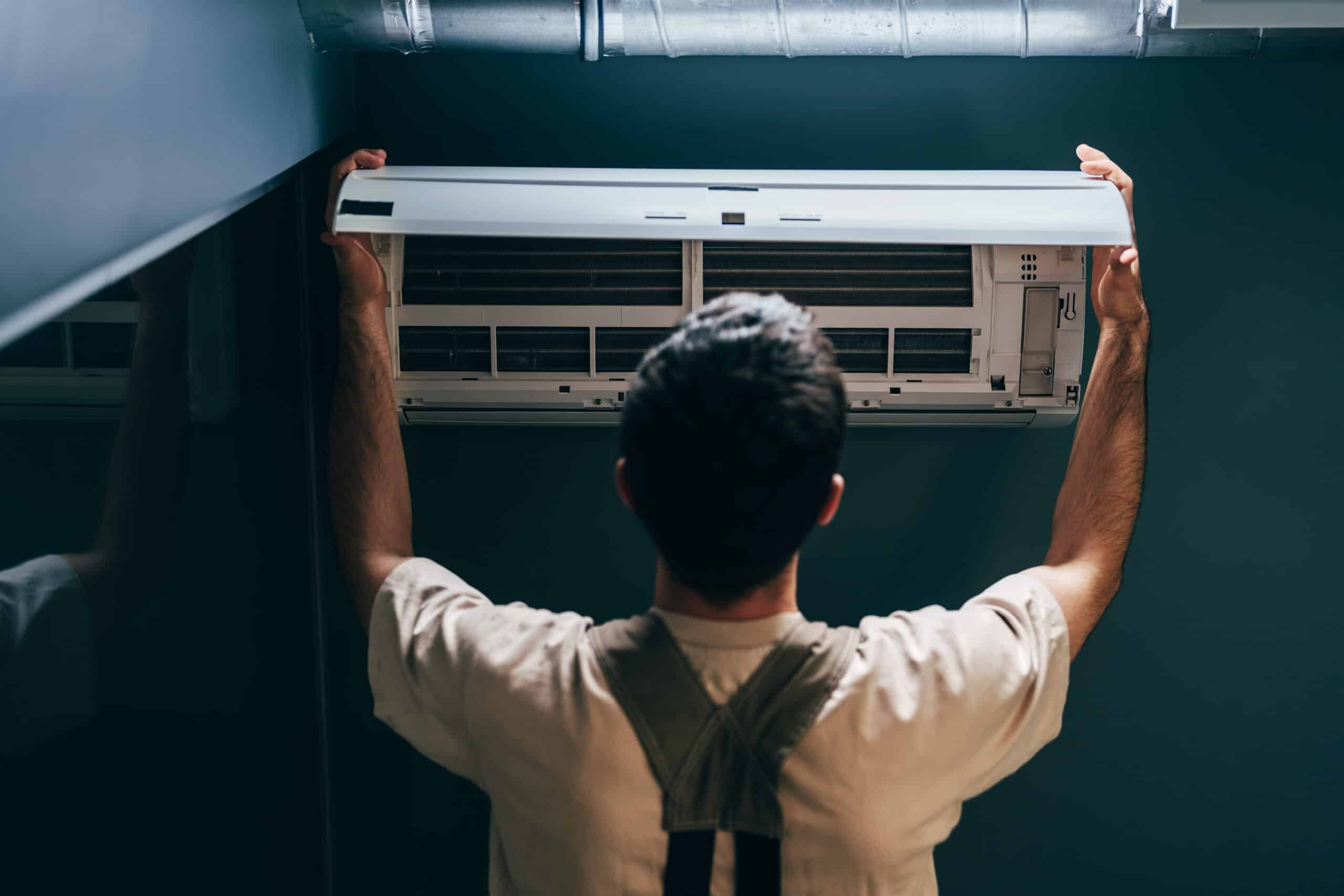 A technician in a light-colored shirt and suspenders is installing or servicing an air conditioning unit mounted on a wall. The setting has a dark background and there is a ventilation duct visible above the unit.