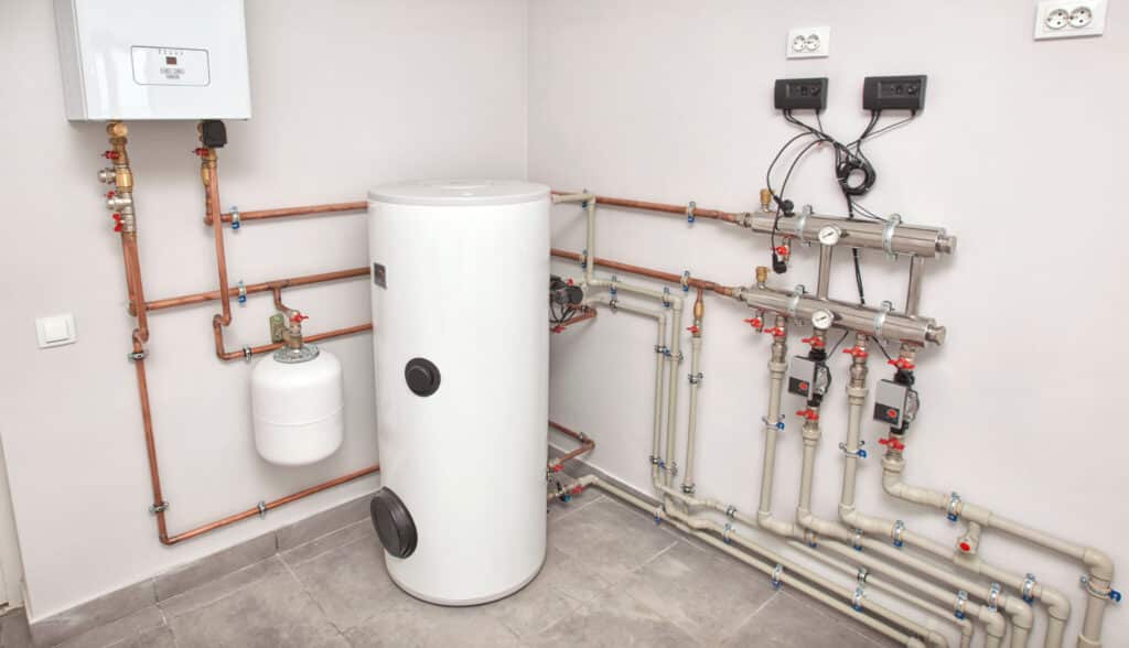 A utility room featuring a white water heater with a cylindrical shape, surrounded by an array of copper and plastic pipes. The walls are light-colored, and two control panels are mounted at the top right, with various valves and connectors visible on the pipes.